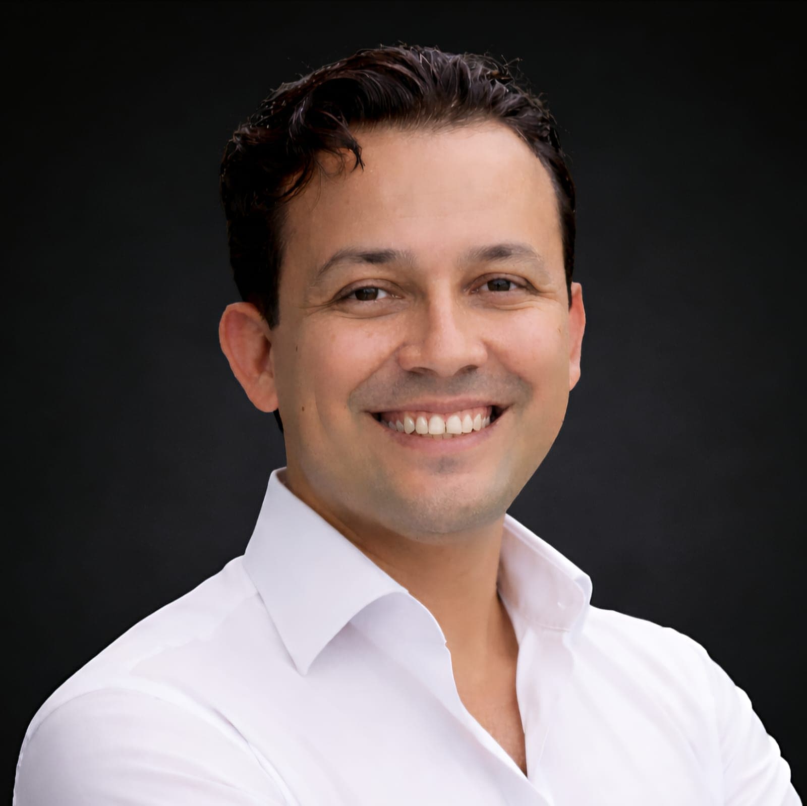 Retrato de un hombre sonriente con camisa blanca y cabello oscuro sobre fondo negro.